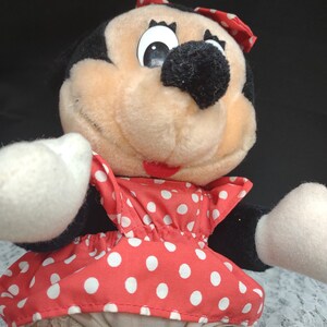 Op de afbeelding: Een pluche speelgoed van Minnie Mouse die een rode jurk met witte stippen en een witte strik draagt. Ze heeft zwarte ogen, een zwarte neus en een roze mond.