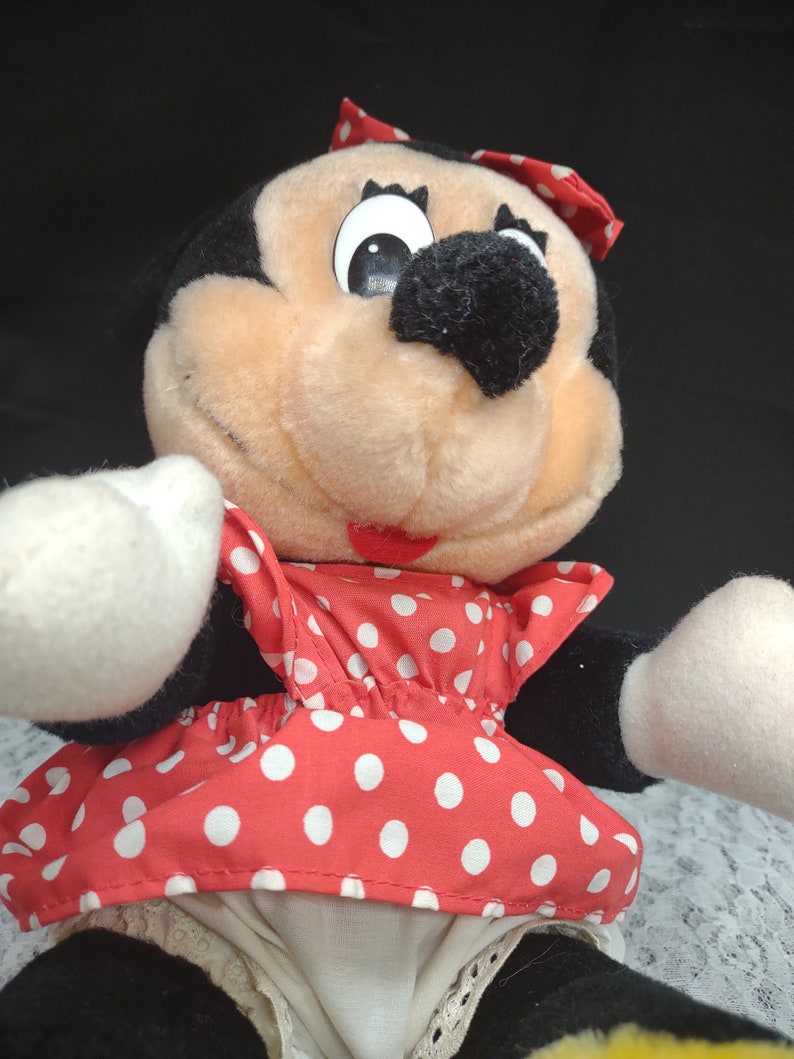 Op de afbeelding: Een pluche speelgoed van Minnie Mouse die een rode jurk met witte stippen en een witte strik draagt. Ze heeft zwarte ogen, een zwarte neus en een roze mond.