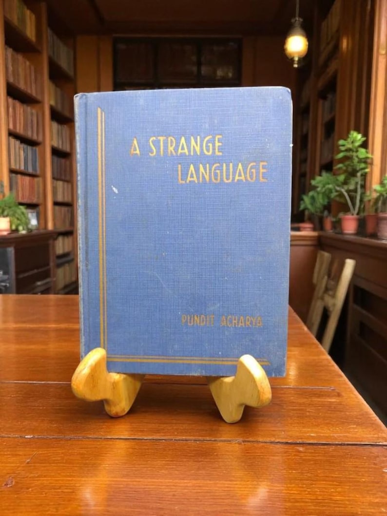 May include: A blue book titled "A Strange Language" with gold lettering, resting on a wooden book stand. The book is displayed on a wooden table in a library setting. The author's name, Pundit Acharya, is printed at the bottom.