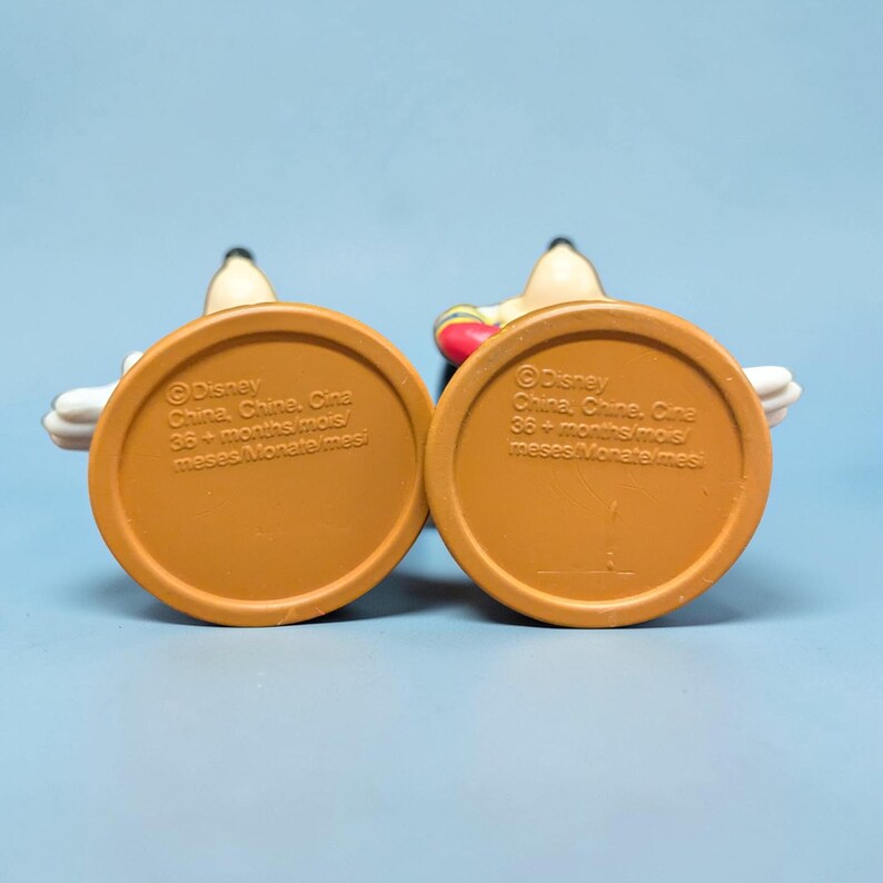 Op de afbeelding: Twee Mickey Mouse-beeldjes, van onderaf gezien, met bruine, cirkelvormige bases. De tekst op de bases luidt "Disney China, Chine, Cina 36+ maanden mois/ meses Monate mesi". De beeldjes staan tegen een lichtblauwe achtergrond.