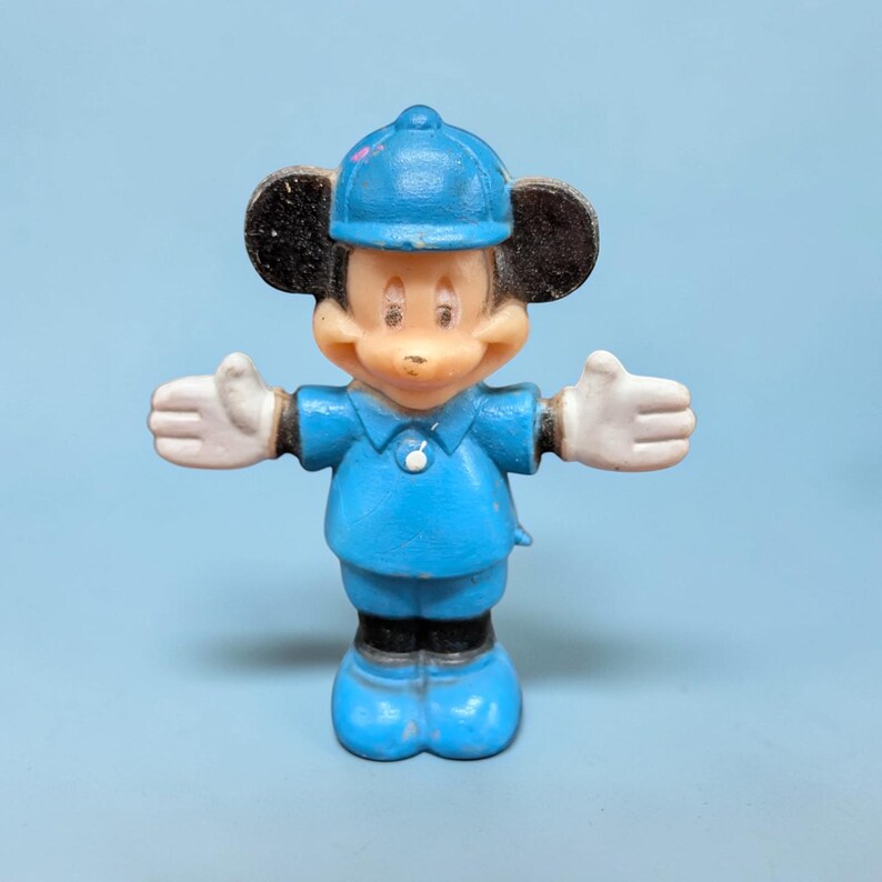 Op de afbeelding: Een vintage Mickey Mouse-beeldje in een blauw uniform en hoed, met witte handschoenen en zwarte oren. Het beeldje staat met uitgestrekte armen op een lichtblauwe achtergrond. Het speelgoed is van plastic.
