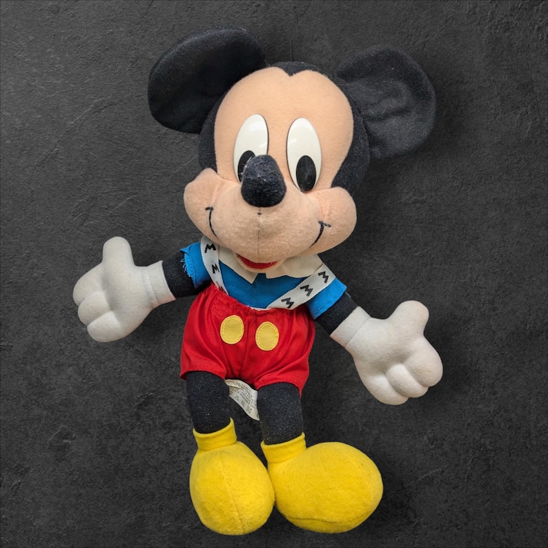 Pu&ograve; includere: Un giocattolo di peluche vintage di Topolino che indossa una camicia a righe blu e bianche con bretelle e pantaloncini rossi. Il giocattolo ha orecchie nere, un naso nero e scarpe gialle.