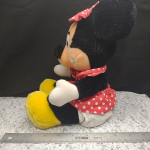 Op de afbeelding: Een pluche speelgoed van Minnie Mouse die een rood-wit geblokt jurkje draagt met een rode strik. Ze heeft gele schoenen en zit op een wit kant.