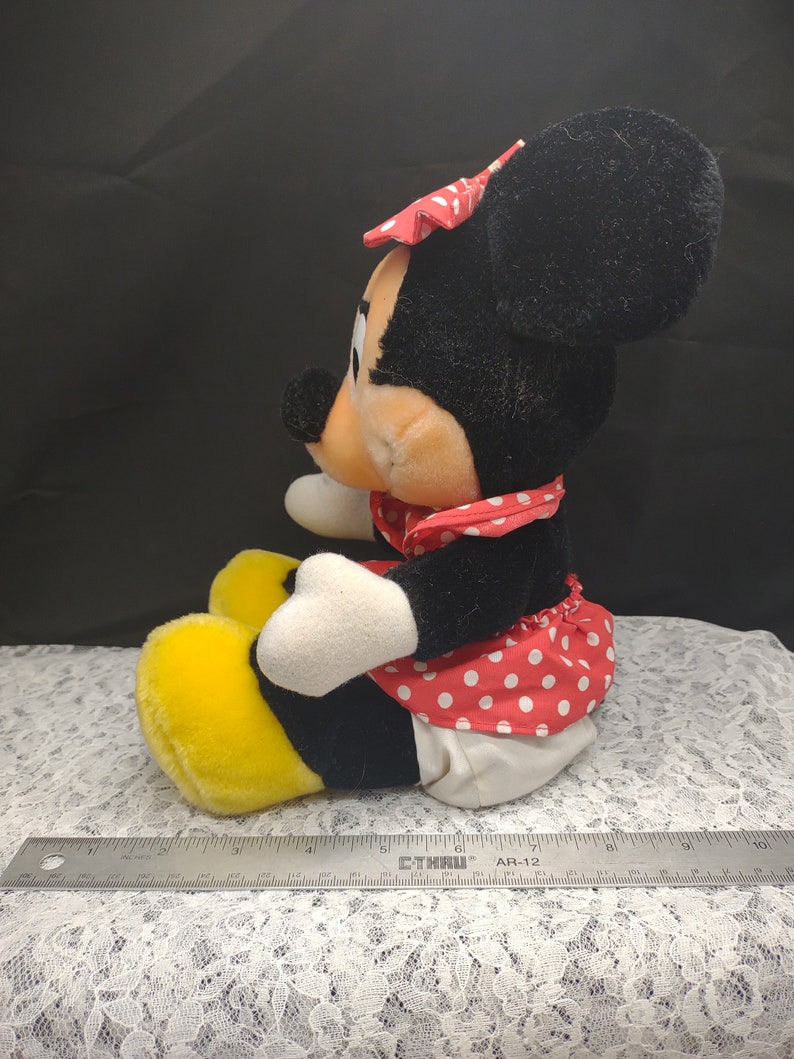 Op de afbeelding: Een pluche speelgoed van Minnie Mouse die een rood-wit geblokt jurkje draagt met een rode strik. Ze heeft gele schoenen en zit op een wit kant.