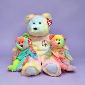 May include: Three colorful plush teddy bears with rainbow-colored fur. Each bear has a peace sign on its chest and a red tag. The bears are in shades of yellow, pink, green, and blue, set against a purple background.