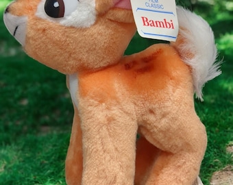 Puede incluir: Un juguete de peluche de Bambi, un cervatillo con cola blanca y ojos marrones. El juguete está de pie sobre una superficie de hierba verde. La etiqueta adjunta al juguete dice "Walt Disney's Animated Film Classic Bambi".
