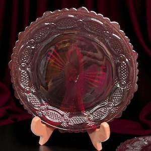 May include: A ruby red glass plate with a scalloped edge and embossed floral and oval patterns. The plate is displayed on a small, light brown stand. The background features a dark red curtain.