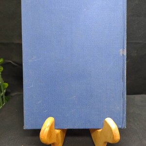 May include: A blue hardcover book with a textured cover is displayed on a wooden book stand. The book's spine is visible, and the stand is made of light-colored wood. The background is dark.