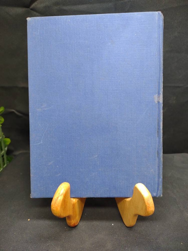 May include: A blue hardcover book with a textured cover is displayed on a wooden book stand. The book's spine is visible, and the stand is made of light-colored wood. The background is dark.