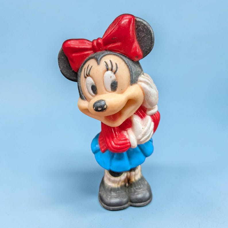 Op de afbeelding: Een vintage Minnie Mouse beeldje. De figuur heeft een rode strik, een rood-witte sjaal, een blauwe rok en zwarte schoenen. Het beeldje is gemaakt van hard plastic en staat op een lichtblauwe ondergrond.