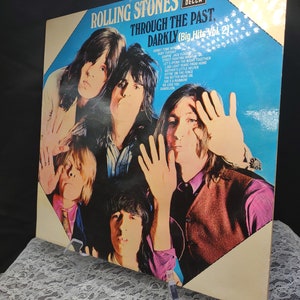 May include: A vintage vinyl record album titled "Through the Past, Darkly (Big Hits Vol. 2)" by The Rolling Stones. The album cover features a group photo of the band members with a blue and white background. The album is displayed on a clear stand.