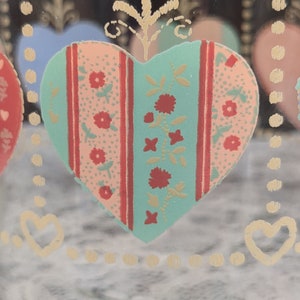 May include: A glass jar with a heart design. The heart is decorated with a pink, red, and green floral pattern. The jar has a gold rim and a gold heart design around the top.
