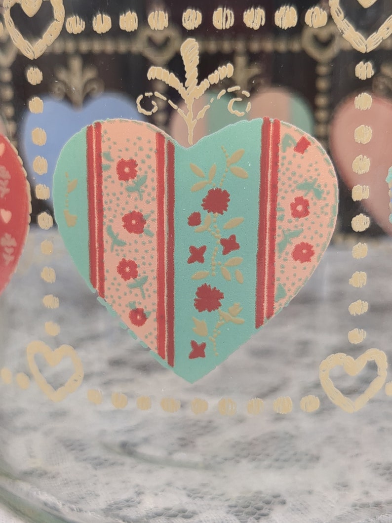 May include: A glass jar with a heart design. The heart is decorated with a pink, red, and green floral pattern. The jar has a gold rim and a gold heart design around the top.