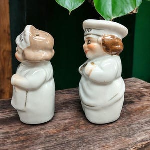 May include: A set of two ceramic salt and pepper shakers shaped like chefs. The salt shaker has the word "SALT" on the hat and the pepper shaker has the word "PEP" on the hat. Both shakers are white with brown hair and faces.