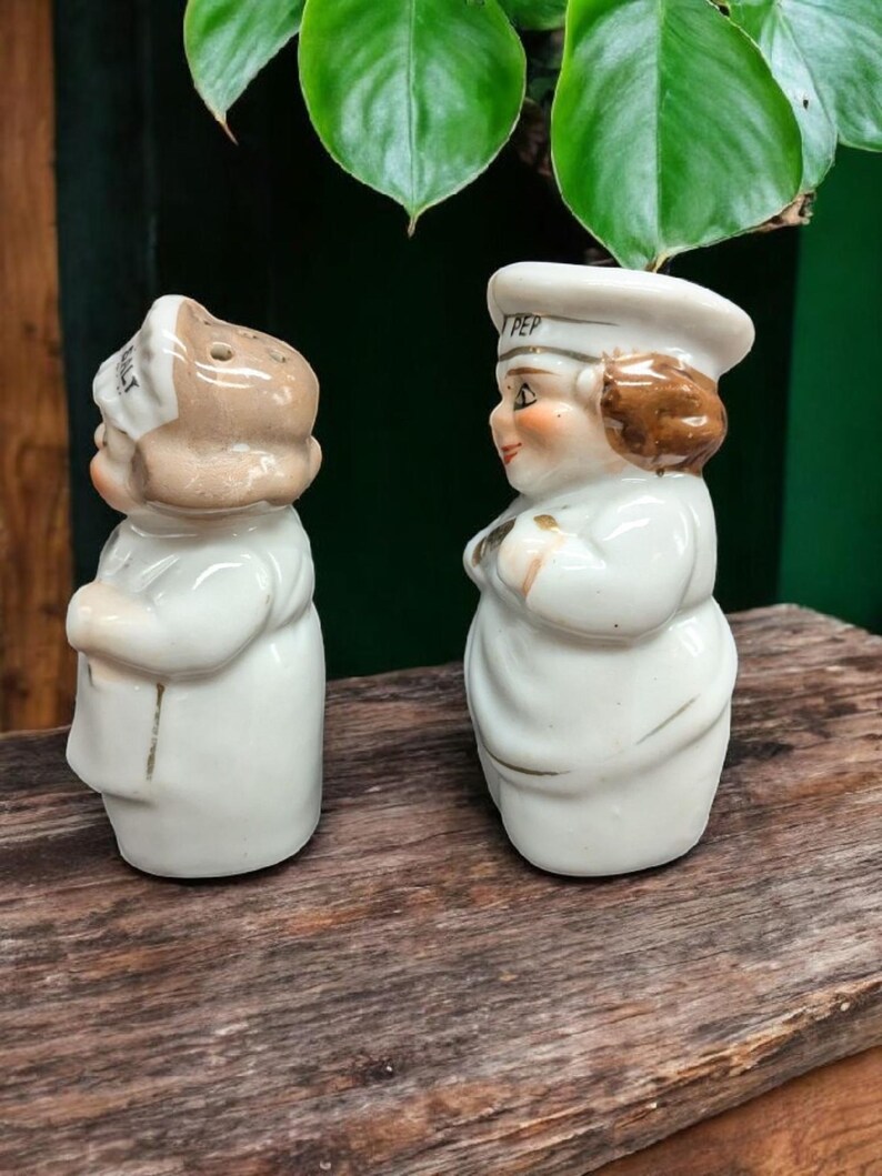 May include: A set of two ceramic salt and pepper shakers shaped like chefs. The salt shaker has the word "SALT" on the hat and the pepper shaker has the word "PEP" on the hat. Both shakers are white with brown hair and faces.