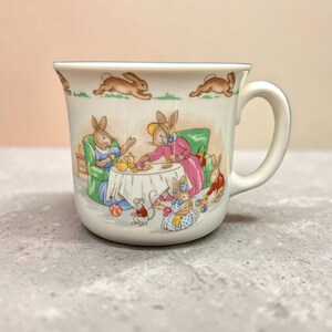 May include: A ceramic mug with a whimsical illustration of rabbits having tea. The mug features a white background with a light blue rim and handle. The illustration depicts rabbits in dresses and suits, seated around a table, with a mouse and toys.