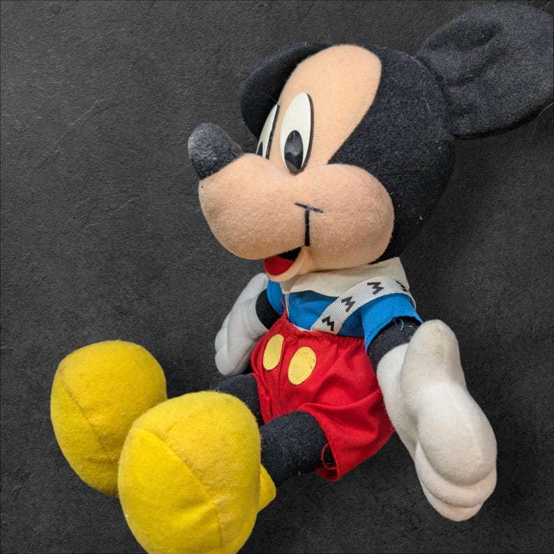 Pu&ograve; includere: Un giocattolo di peluche di Topolino che indossa una camicia blu e bianca con una salopette rossa e scarpe gialle. Il giocattolo ha un naso nero e grandi orecchie nere.