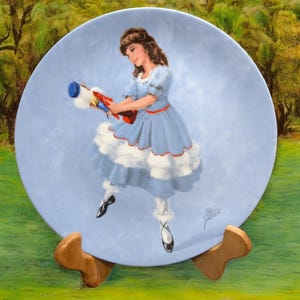May include: A blue and white porcelain plate with a painting of a young girl in a blue dress holding a nutcracker. The plate is on a wooden stand.