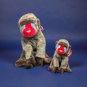 May include: Two plush mandrill monkeys. The larger one is seated, and the smaller one is in front. Both have brown and gray fur, red noses, and blue markings. Each has a red tag.