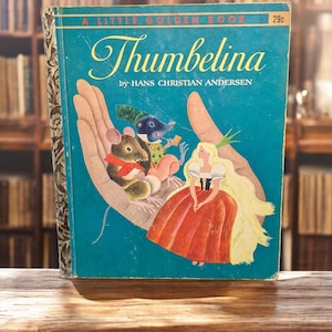 May include: A Little Golden Book titled "Thumbelina" by Hans Christian Andersen. The cover is teal with illustrations of a blonde woman in a red dress, mice, and a hand. The book is on a wooden surface.