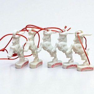 May include: A row of five white goat figurines with red string attached. Each figurine stands on a rectangular base with the word "Celebrator" printed in red. The goats are in a standing pose.
