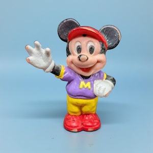 Op de afbeelding: Een vintage Mickey Mouse beeldje in een paars shirt met een gele "M", gele shorts en rode schoenen. Het beeldje heeft een rode pet en witte handschoenen, met zwarte oren en een lachend gezicht. De achtergrond is effen lichtblauw.