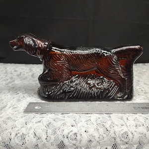 May include: A brown glass bottle shaped like a dog, possibly a bloodhound, with a stopper. The bottle has a detailed design of the dog's fur and features.