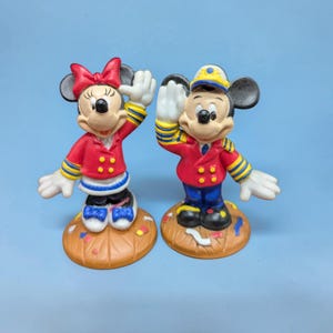 Op de afbeelding: Twee keramische beeldjes van Mickey en Minnie Mouse in dirigentenoutfits. Mickey draagt een rode jas, een blauwe broek en een geel omrande hoed. Minnie draagt een rode jas en een rode strik. Beide staan op bruine voetstukken.