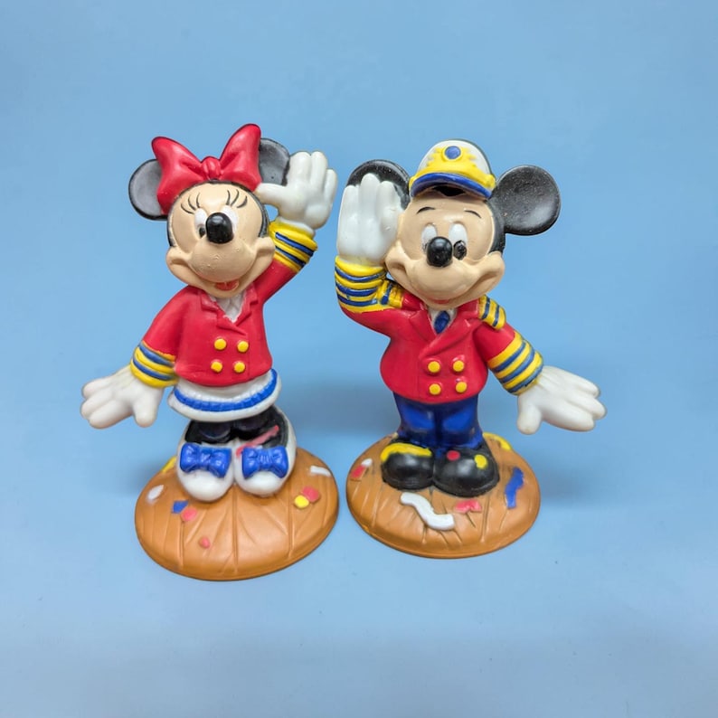 Op de afbeelding: Twee keramische beeldjes van Mickey en Minnie Mouse in dirigentenoutfits. Mickey draagt een rode jas, een blauwe broek en een geel omrande hoed. Minnie draagt een rode jas en een rode strik. Beide staan op bruine voetstukken.
