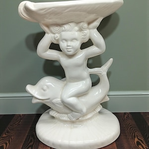 May include: A white ceramic pedestal table featuring a cherub riding a dolphin, supporting a large shell-shaped bowl. The base is round with a textured, scalloped design. The sculpture is a decorative accent piece.