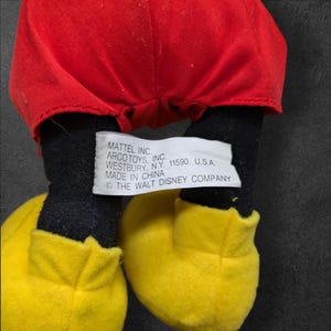 Pu&ograve; includere: Un giocattolo di peluche Mickey Mouse nero e rosso con scarpe gialle. Il giocattolo ha un'etichetta sul fondo che recita "MATTEL INC. ARCOTOYS, INC. WESTBURY, N.Y. 11590. U.S.A. MADE IN CHINA &copy; THE WALT DISNEY COMPANY"