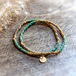 Könnte beinhalten: Ein doppelreihiges Armband mit türkisfarbenen und goldenen Perlen, mit einem kleinen goldenen Anhänger mit Blumenmuster.