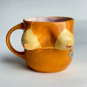 Könnte beinhalten: Eine Keramik-Tasse in einem warmen Orangeton, mit einem einzigartigen Design einer Büste mit cremefarbenen, abgerundeten Brüsten. Die Tasse hat einen kleinen goldenen Anhänger mit einem rautenförmigen Anhänger, der an einer der Brüste hängt. Das Innere der Tasse ist lila und rosa.