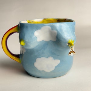 Può includere: Una tazza in ceramica blu con un manico arcobaleno e nuvole bianche dipinte sul davanti. Un piccolo ciondolo dorato con un design arcobaleno pende dalla tazza.