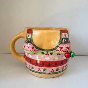 May include: A ceramic mug shaped like a bust wearing a festive Christmas sweater with red, green, and white stripes, snowflakes, and candy canes. The mug has two small green and red jingle bells hanging from the sweater.