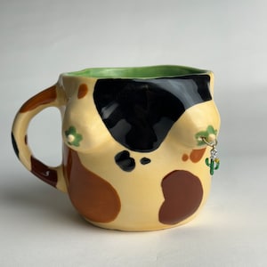 May include: A ceramic mug with a brown, black and green abstract design. The mug has a handle and a small cactus charm hanging from a ring on the side.