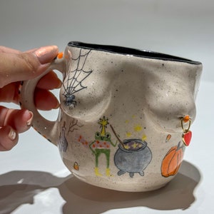 Puede incluir: Taza de cerámica beige con borde negro, con ilustraciones pintadas a mano con temática de Halloween. La taza tiene una telaraña y una araña, una rana con una varita, un caldero y una calabaza. Un pendiente dorado con un dije de corazón rojo.