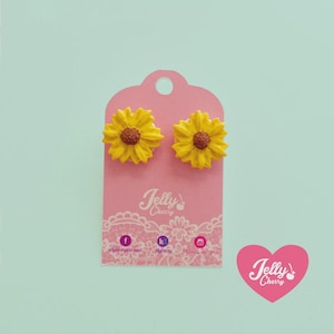 Puede incluir: Un par de pendientes de botón con forma de girasoles amarillos y centros marrones, montados sobre una tarjeta de presentación rosa con el texto Jelly Cherry y patrones de encaje blanco, junto a un logo de corazón rosa con Jelly Cherry.
