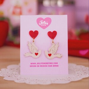 Puede incluir: Un par de pendientes en una tarjeta de presentación rosa claro con el texto Jelly Cherry y www.jellycherrymx.com Hecho en Mexico con amor. Los pendientes consisten en un poste de corazón rojo unido a una paloma color crema con un detalle.
