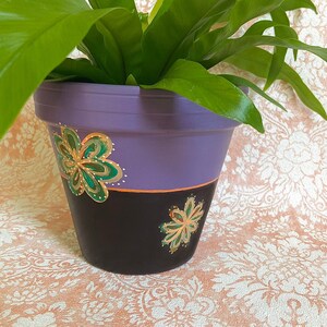 May include: A purple and black terracotta planter with green and gold painted flowers. The planter is filled with a green leafy plant.