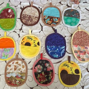 May include: A collection of eleven oval-shaped fabric pot holders, each featuring a hanging loop at the top. The pot holders display various colorful patterns, including sunflowers, turkeys, cacti, butterflies, autumn leaves, and holiday-themed prints.