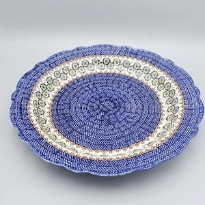 May include: A round ceramic plate with a scalloped edge featuring a deep blue center covered in small white dots. The border displays a repeating pattern of yellow and white flowers with green leaves on a cream background, framed by blue.