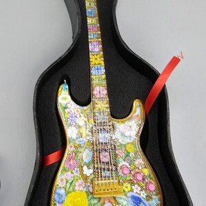 May include: A miniature electric guitar with a floral design painted on the body and neck. The guitar is in a black case with red ribbon.