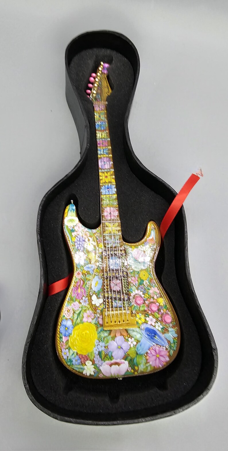 May include: A miniature electric guitar with a floral design painted on the body and neck. The guitar is in a black case with red ribbon.