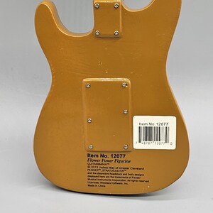 May include: A gold-colored electric guitar figurine with the text "Item No. 12077 Flower Power Figurine GUITARMANIA 2013 United Way of Greater Cleveland FENDER STRATOCASTER and the distinctive headstock and body designs displayed here are the trademarks of Fender Musical Instruments Corporation. All rights reserved. Licensee: Westland Giftware, Inc. Made in China" on the back.