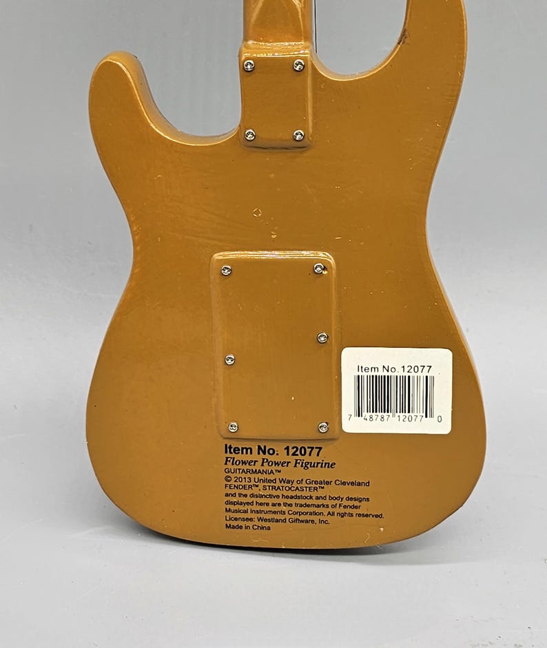 May include: A gold-colored electric guitar figurine with the text "Item No. 12077 Flower Power Figurine GUITARMANIA 2013 United Way of Greater Cleveland FENDER STRATOCASTER and the distinctive headstock and body designs displayed here are the trademarks of Fender Musical Instruments Corporation. All rights reserved. Licensee: Westland Giftware, Inc. Made in China" on the back.