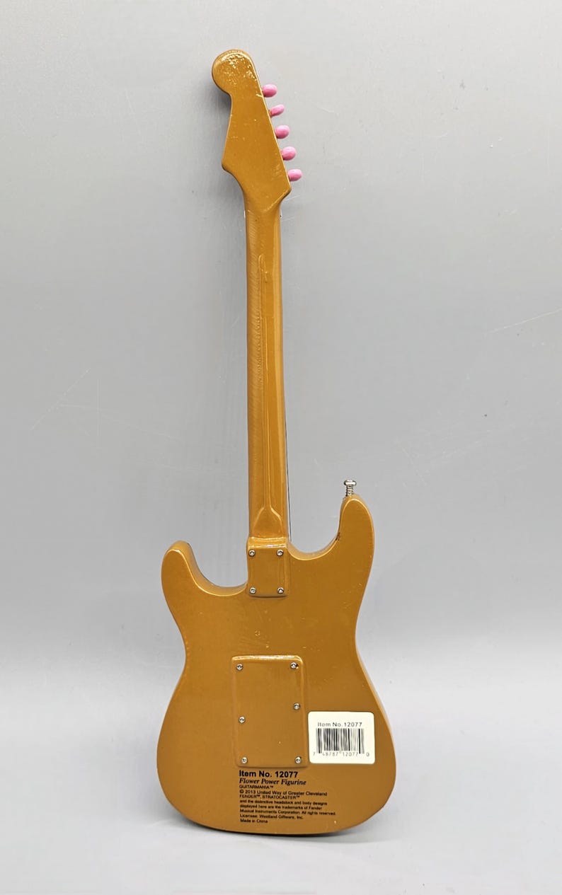 May include: Gold-colored electric guitar figurine with pink tuning pegs. The back of the figurine has a label with the text "Item No. 12077 Flower Power Figurine" and a barcode.