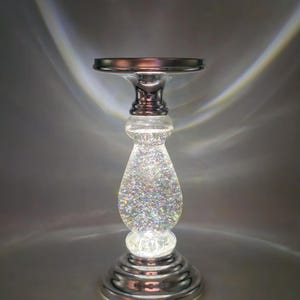 May include: A decorative candle holder with a silver-colored base and top. The central portion is a clear, teardrop-shaped glass filled with sparkling glitter. The holder is illuminated from within, creating a shimmering effect.