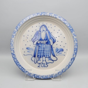 May include: A white ceramic plate with a blue rim and a blue Santa Claus design. The Santa Claus figure is wearing a blue robe and holding a staff. The year "2015" is printed below the figure. The plate is on a clear stand.