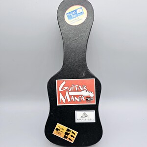 May include: Black guitar-shaped box with stickers. The stickers include "Guitar Mania", "Rock 'n' Roll", "Fender", and a United Way logo.